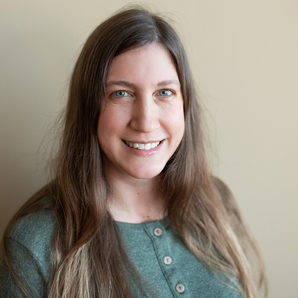 White woman with long brown hair wearing a light green shirt with three buttons down the middle stands in front of a beige wall smiling and posing for the camera.