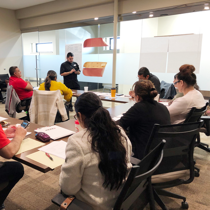 A group of adults sit around a conference table in a modern meeting room, listening to a man standing at the front giving a presentation. Some participants are taking notes while others look at their phones. A flip chart and papers are visible in the background.