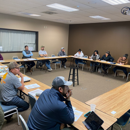 A group of people sit around conference tables that are arranged in a square. Everyone has papers in front of them and they appear to be listening to a speaker.