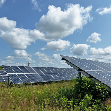 3 paneles solares con pastos de pradera debajo. Un cielo azul con nubes esponjosas arriba.