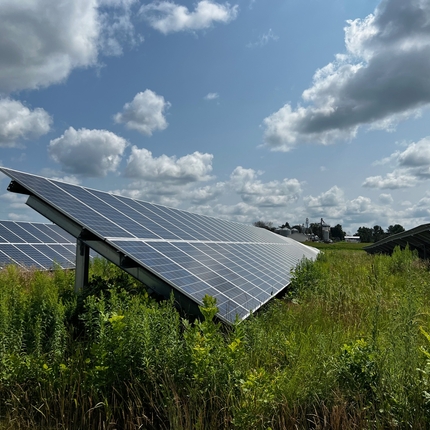 Dos paneles solares largos orientados hacia la derecha de la cámara, con cultivos debajo. El cielo es azul y tiene nubes esponjosas.