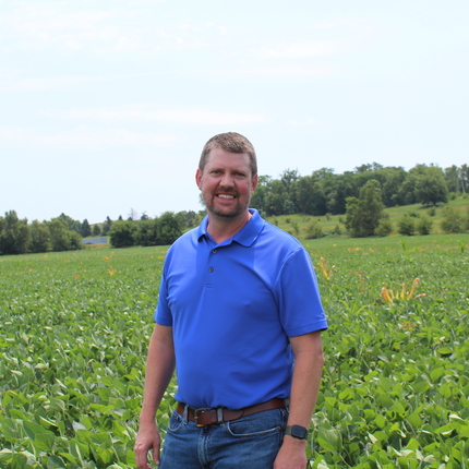 Man with short hair and facial hair smiles at the camera. He is standing in the middle of a soybean field. The beans are leafy green with purple flowers. The man is wearing a collared short sleeve blue shirt.