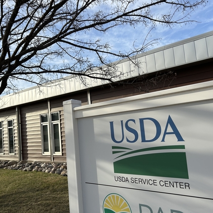 Centro de Servicio del USDA: la foto muestra la mitad superior de un letrero con el logotipo verde y fondo blanco. Detrás del letrero se encuentra un edificio de una planta con revestimiento marrón, con las ramas de un árbol con brotes primaverales extendiéndose hacia el edificio y un cielo azul con nubes tenues.