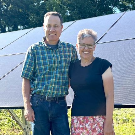 Un hombre y una mujer están de pie, abrazados, frente a un conjunto de paneles solares. Se encuentran sobre un césped de un verde brillante, con árboles de hojas verdes al fondo.