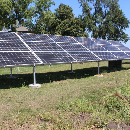 Un panel solar, con césped verde debajo y una hilera de pinos más un contenedor de granos detrás.
