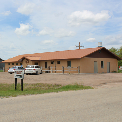 A tan, one-story commercial building with a reddish-brown metal roof and wooden fence sits beside a gravel parking lot. Two white vehicles are parked in front of the building, which has a round "C" logo above the entrance. A sign in the foreground reads "C Market Entrance." The sky is mostly clear with scattered clouds, and a water tower labeled "CODY" is visible in the background.