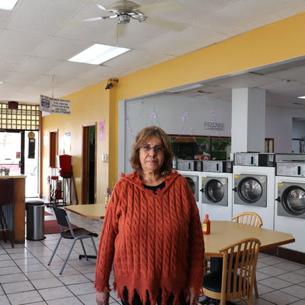 Hispanic woman with short brown hair, glasses, and an orange long sleeve sweater stands in the center of a laundry business with washing machines and brown tables with chairs behind her