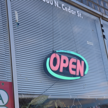 Letrero eléctrico que dice "open" en una ventana de vidrio enfrente de persianas blancas