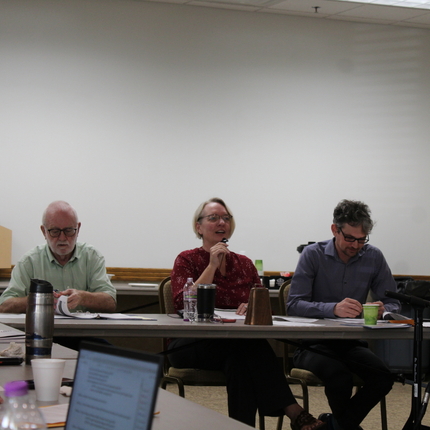Three white individuals, including two men and a woman sit at a long table, woman has a microphone in hand, while a hispanic woman stands in the corner of the room