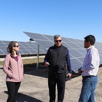 Tres personas están de pie en un campo. Detrás de ellas hay hileras de paneles solares y las personas conversan sobre conservación.
