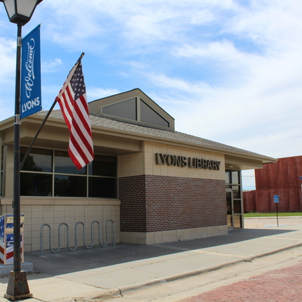 A brick and tan building with large windows that says “Lyons Library.” In front of the library are lamp posts with blue banners that say “Welcome to Lyons” and which have American flags. Also in front of the building is a bike rack and a flag drop-off receptacle along a sidewalk.