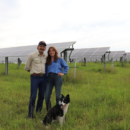Man and woman standing in a field of longer grass. A black and white dog is in front of them. Solar panels are behind them. 