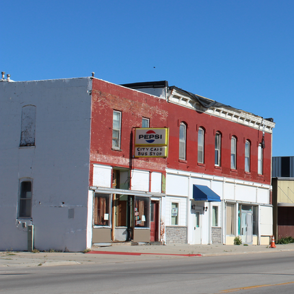 An old, two-story brick building sits along a highway. It appears to be vacant. On the upper half, we see red brick and a line of windows. The ground level has painted white brick. There is an old sign that says "PEPSI CITY CAFE BUS STOP."