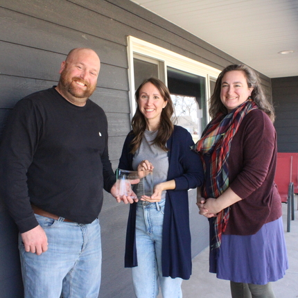 Woman wearing blue sweater and jeans presents an award to a man wearing a black shirt and jeans. A woman wearing a maroon shirt and blue skirt stands on the right.
