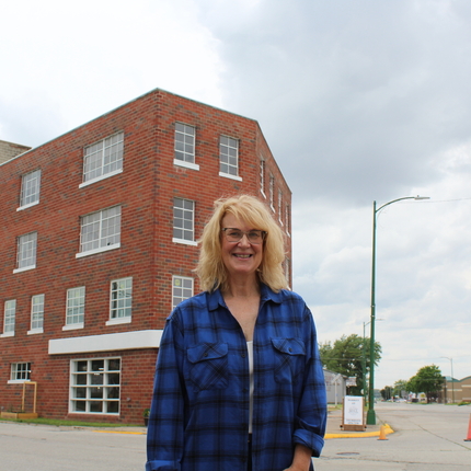 Woman wearing a blue plaid button up shirt, with glasses and blond shoulder-length hair, standing in front of a 4 story old building (a mill) with cloudy skies