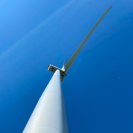 Primer plano de un aerogenerador blanco, mirando hacia el motor y las hélices. El cielo es azul brillante al fondo. 