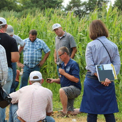 Photo of people standing and kneeling in front of a green corn field. They're looking  at the hand of a man holding a microphone in one hand and soil in the other. 