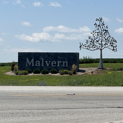 Letrero en la esquina de dos carreteras, colocado en el suelo con pequeñas plantas ornamentales a su alrededor. Dice "Malvern". A la derecha del letrero hay una obra de arte de metal con forma de árbol.