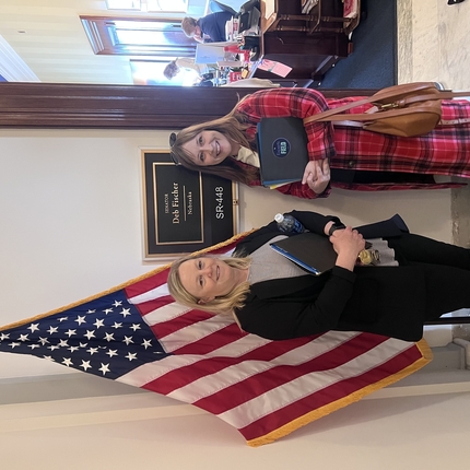 Dos dones de pie frente a un lletre, fora d'una oficina, que dice «Deb Fischer Nebraska», junt a una bandera nord-americana.