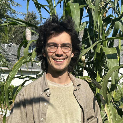 Man with shoulder length black hair and glasses wearing light green pants and shirt, with a long sleeve tan shirt over, stands in front of some corn 