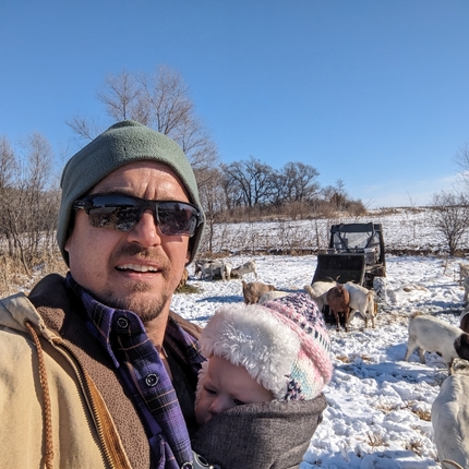 Un hombre blanco en un abrigo color cafe y gorro verde carga a un  bebe cubierto en una cobija color oscuro con una gorra color blanco y rosa. A su espalda esta un campo cubierto en nieve y cabras comiendo de heno que puso él.