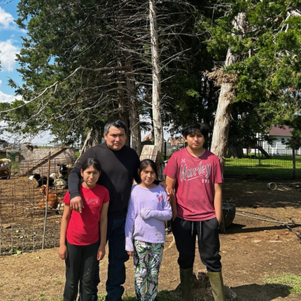 Latino family with two girls, one boy, and a dad, poses in front of a pen with goats and pine trees 