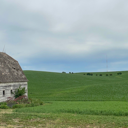 Old white barn sits on plot of green land with electrical windmill in far right corner