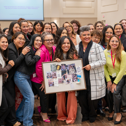 Picture of attendees at the 2024 Latina Women in Business Conference. They are all smiling, huddled around a young Latina women holding a vision board. She has long, dark hair, and smiles at the camera. 