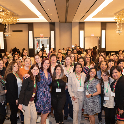 Over 20 women pose por a picture in a conference hall with gold chandeliers above and tables around