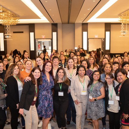Más de 20 mujeres posan para una foto en una sala de conferencia con mesas redondas a la orilla de la foto