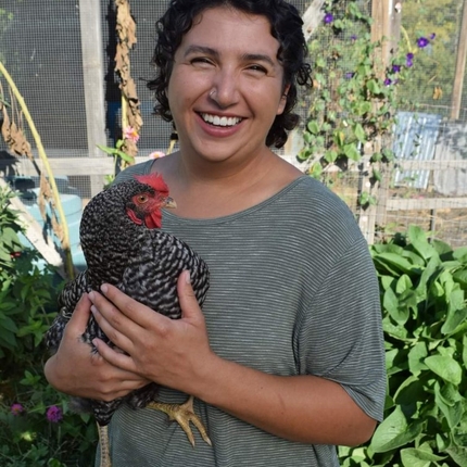 Mujer de cabello oscuro, sonriendo a la cámara, con una camisa gris de manga corta, sosteniendo un pollo. Al fondo, una dependencia con plantas.
