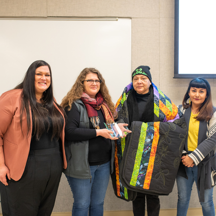 Four women smiling at the camera, two holding an award, one holding a Native star quilt