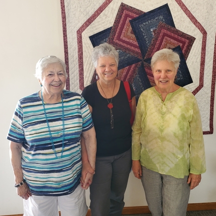 Tres mujeres mayores de pie delante de una colcha hecha a mano.
