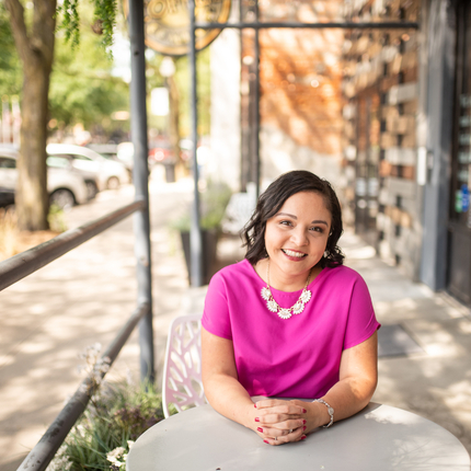Woman smiling with short dark hair sitting at small round table wearing a bright pink shirt holding both hands together