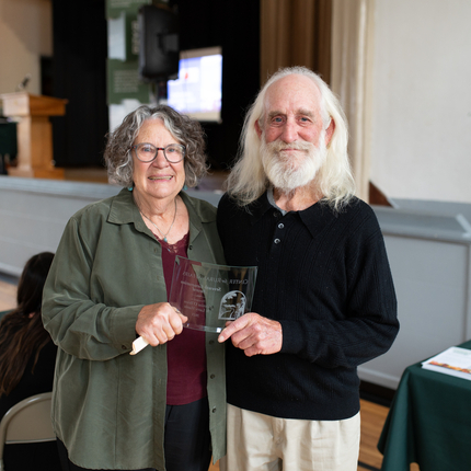 Woman and man holding a clear freestanding award plaque. Woman has short curly gray hair and is wearing a green long sleeve shirt. Man is wearing longer gray hair with a black long sleeved shirt.