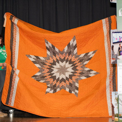 Man with long gray hair and a long sleeved black shirt holds up an orange quilt with a gray star in the center.