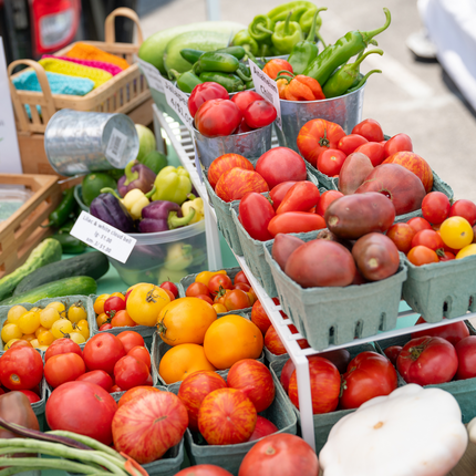 Colorful display of fresh produce at a farmers market, including a variety of heirloom tomatoes in shades of red, yellow, and orange; green bell peppers; chili peppers; and cucumbers. The produce is arranged in tin buckets and green cartons.