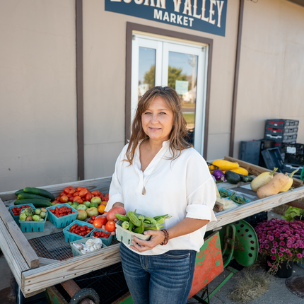 A woman stands in front of a market stand at Logan Valley Market, holding a basket of fresh produce including tomatoes and okra. She is wearing a white blouse and jeans, smiling warmly at the camera. The market table behind her is filled with a colorful variety of vegetables like tomatoes, squash, zucchini, garlic, and eggplant, set against a rustic backdrop.