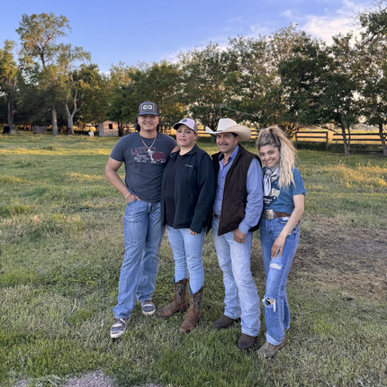 A Latino family, from left, teenage boy, mom, dad, teenage daughter. All are wearing blue jeans and casual clothes, standing in a pasture with trees and a fence behind.