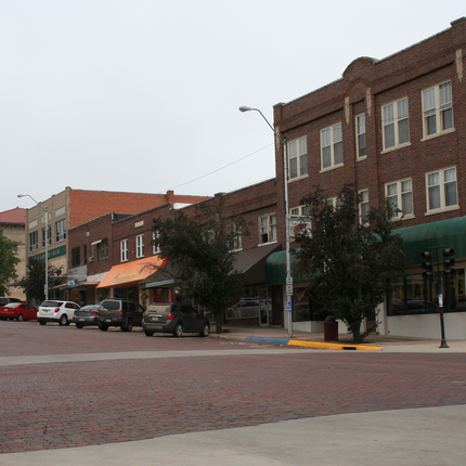 Vista de los edificios de la calle principal de McCook, Nebraska. En el extremo izquierdo, hay un gran edificio de ladrillo claro, junto a él hay un edificio de dos pisos de ladrillo oscuro, y luego otro edificio de dos pisos de ladrillo con un toldo naranja. A la derecha, hay dos edificios más de dos pisos de ladrillo oscuro, cada uno con toldos marrón oscuro. A la derecha de estos edificios hay un edificio de tres pisos de ladrillo oscuro con un toldo verde oscuro. Hay vehículos a lo largo de la calle frente a estos edificios.