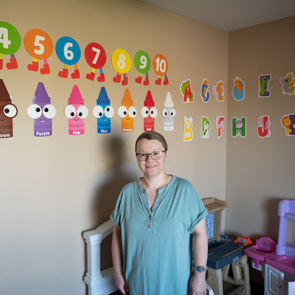 Woman in a short sleeved blue shirt, with pulled back dark hair, and glasses, smiles at the camera. She is standing in front of a beige wall with children toys stacked up behind her. On the wall behind her are colorful numbers, crayons, and letters.
