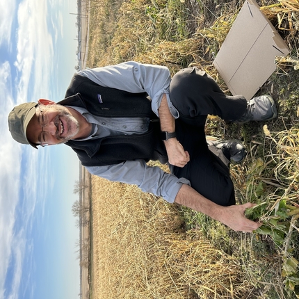 Man crouching in a field with a turnip at his feet. Man is smiling at the camera, wearing a green ball cap, glasses, long-sleeved button up shirt, black vest, and black pants.