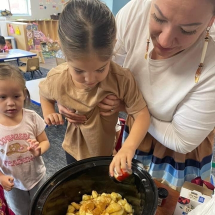 Una mujer sostiene a una niña para que añada ingredientes a una olla de cocción lenta. La olla tiene patatas.