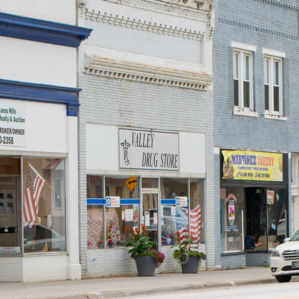Imagen de la fachada de la farmacia Valley Drug Store en Missouri Valley, Iowa. Es un edificio de ladrillo blanco con grandes ventanales y un cartel que dice "Valley Drug Store" y tiene dos plantas grandes en macetas grises que flanquean la puerta. A la derecha hay un edificio de ladrillo azul con un cartel amarillo que dice "Martinez Bakery".