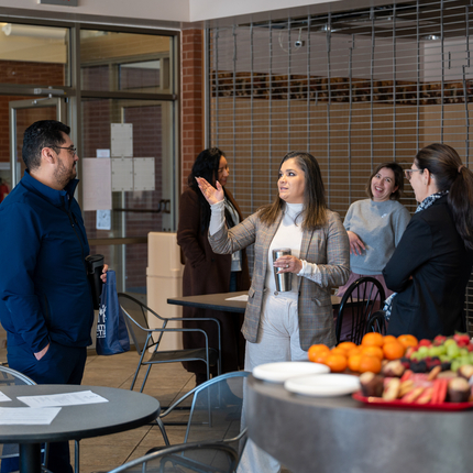 Two clusters of people stand behind two small round tables, with the nearest one full of fruits and pastries. The closest cluster of people has two people. A man is listening to a woman talking and gesturing.