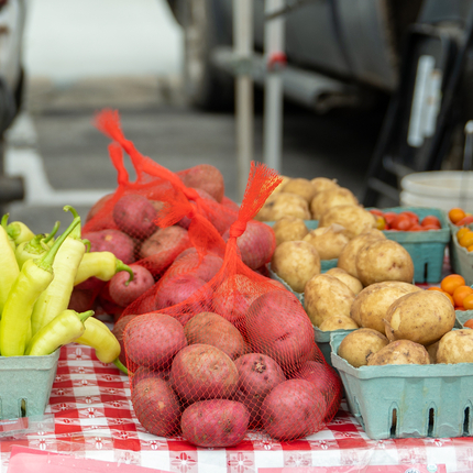 Baskets of vegetables on a table covered with a red and white checkered tablecloth. Vegetables include green cucumbers, light green banana peppers, red potatoes in a red mesh bag, brown small potatoes, cherry tomatoes.