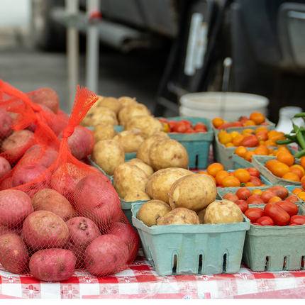 Productos frescos exhibidos en un mercado de agricultores, incluidas papas rojas en bolsas de malla, papas rojizas, tomates cherry y judías verdes dispuestas en cajas de cartón sobre un mantel a cuadros rojos y blancos.