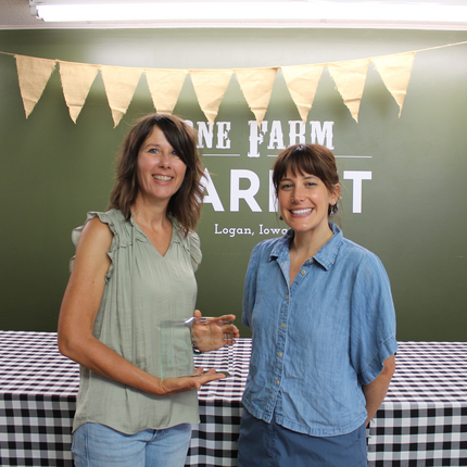 Two women stand side by side, smiling at the camera. The woman on the left has shoulder-length brunette hair, a sage-colored sleeveless top, and holds a rectangular glass award. The woman on the right has brunette hair with bangs, swept back in a ponytail. She wears a short-sleeve chambray button-down shirt. Behind them, there is a table with a black and white plaid table cloth and a green wall that says "One Farm Market Logan, Iowa" with burlap pennants hanging from the ceiling.