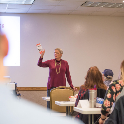 White woman with gray short hair wearing a long sleeve burgundy top holds up a brochure in front of a room of people with their backs towards the camera