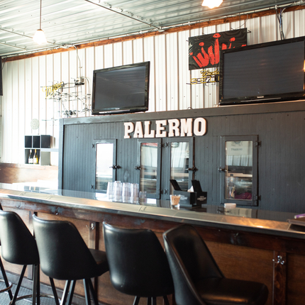 Un bar con taburetes al frente, con "Palermo" en la pared, con dos televisores apagados en la pared encima de la palabra